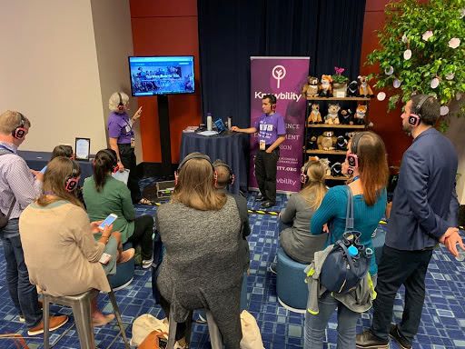 Becky and Antony in purple Knowbility shirts and wearing headphones are showing content on a large screen to audience members who are also wearing hedphones.