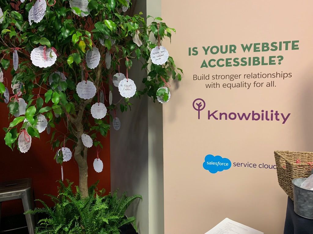 Photo from event showing Knowbility and Salesforce poster as well as a tree with written reflections on small paper flowers hanging from the branches.