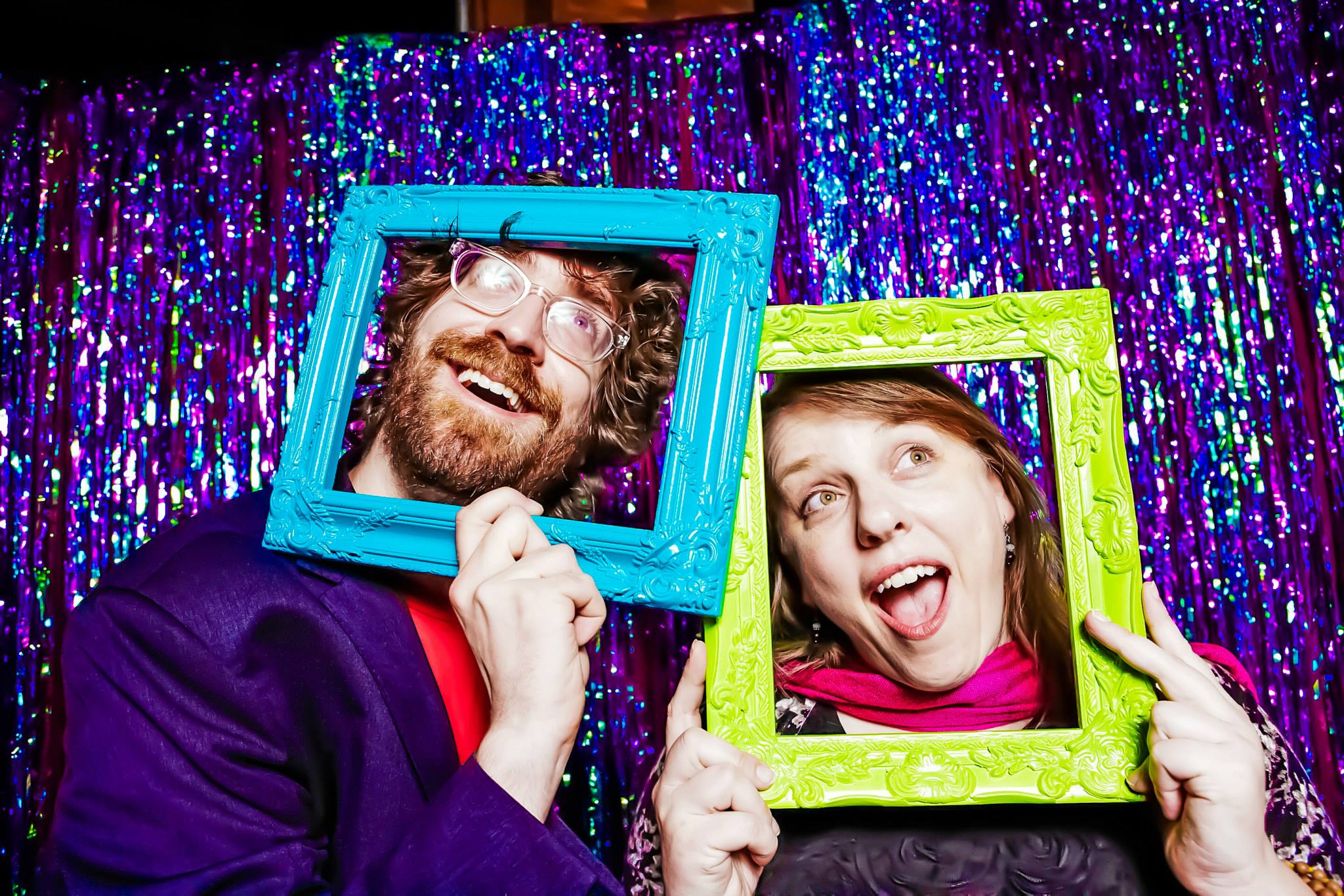Christopher Schmitt and Ari Stiles standing next to each other in front of a glitter background, both with their heads sticking through bright blue and green picture frames, making faces.